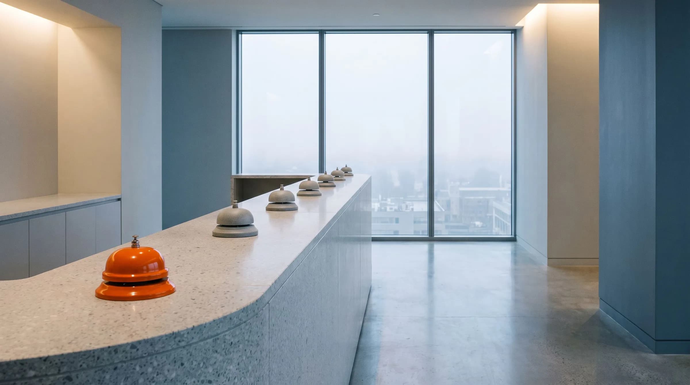 A long terrazzo reception counter with a row of grey service bells and one orange bell at the near end, a foggy city beyond the window