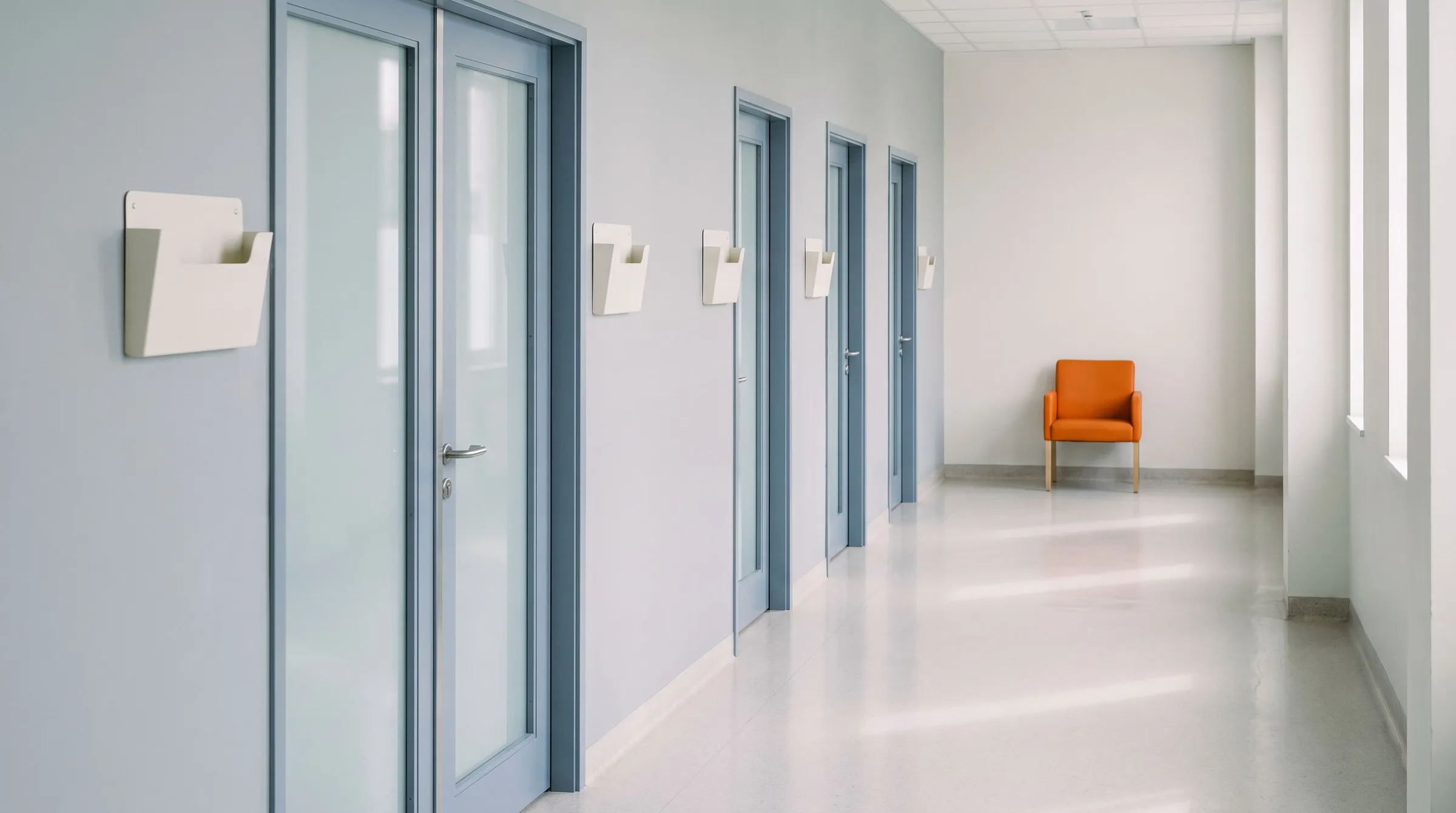 A calm, pale hospital corridor with frosted glass doors and wall chart holders, and a single orange chair at the far end
