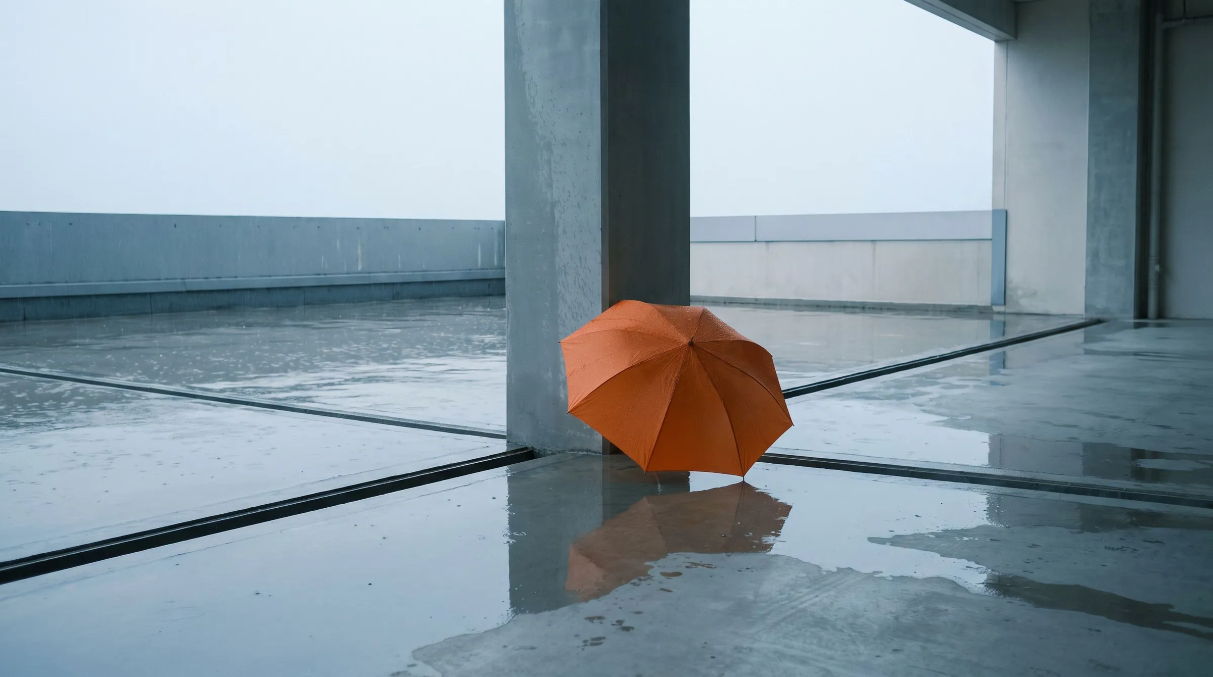 A rain-washed concrete loading court with drainage channels and a single orange umbrella resting against a grey column