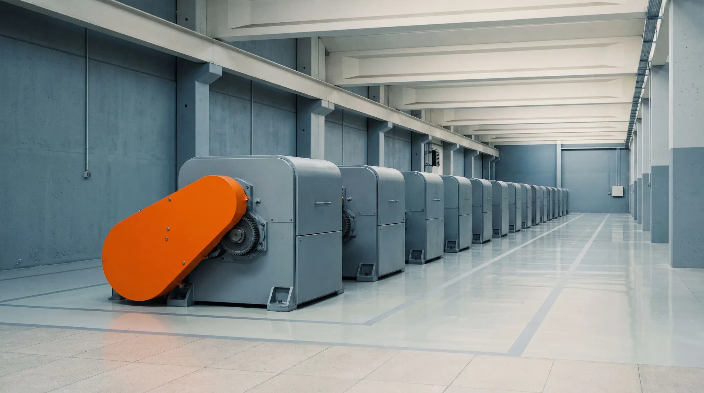 A clean factory hall with a row of identical grey machines and a single orange safety guard on the nearest one