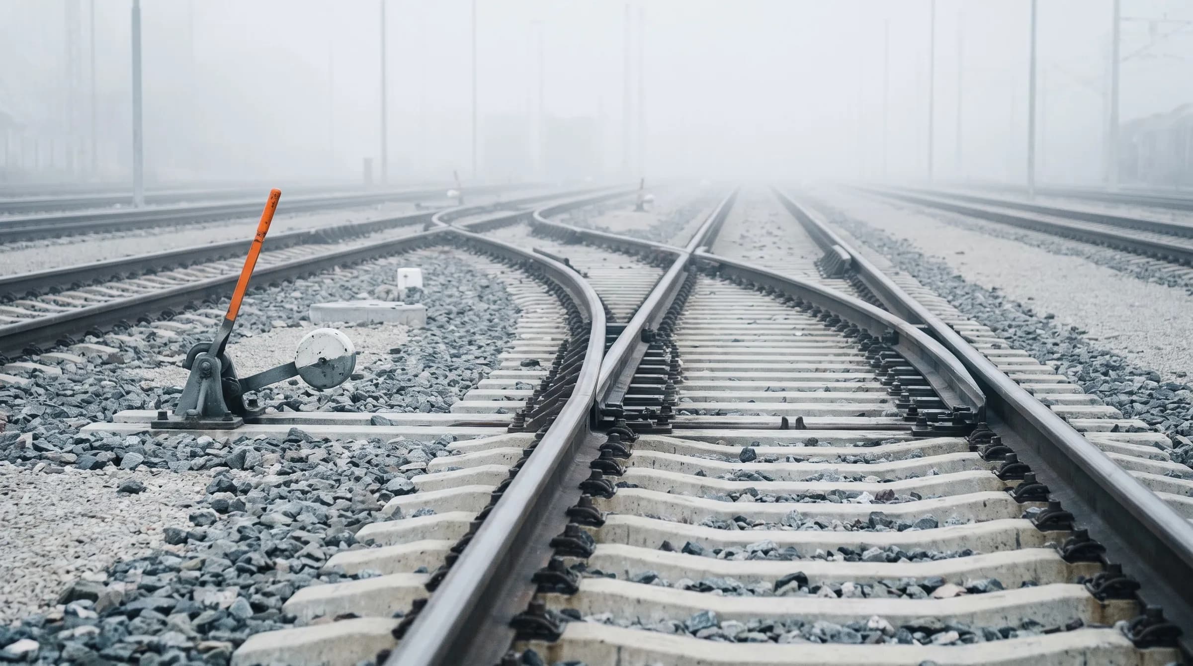 Grey railway tracks in a foggy rail yard where one line curves away at a switch, with a single orange switch lever beside the junction