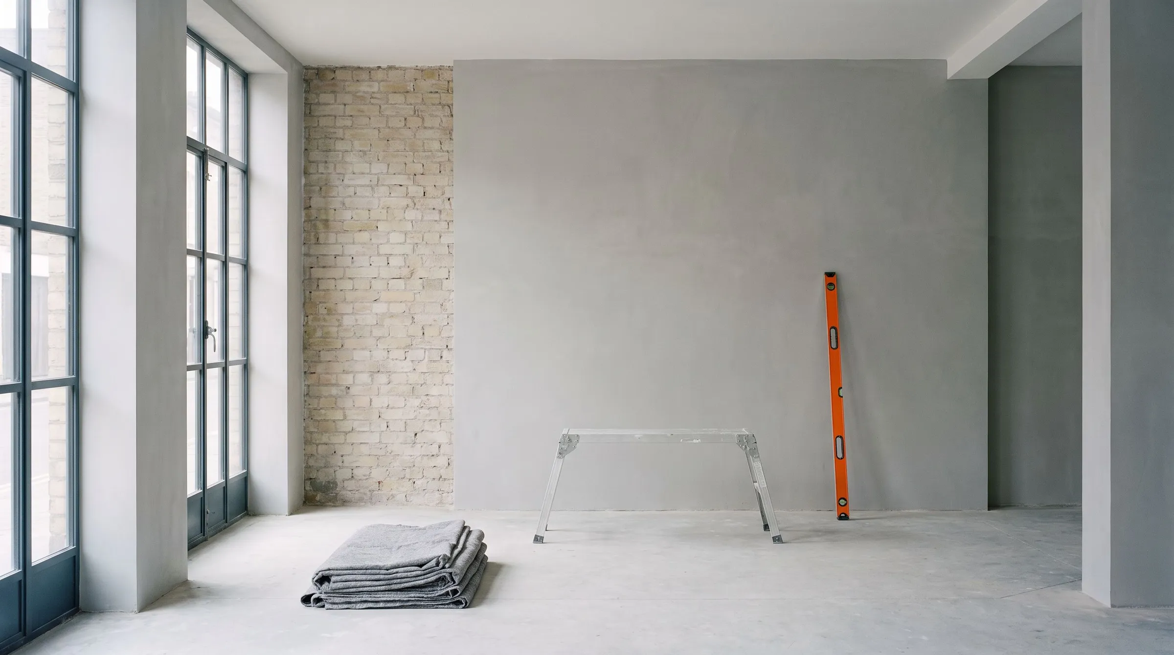 A bright room mid-renovation with steel-framed windows, one old brick wall, folded drop cloths, a trestle and an orange spirit level against fresh plaster