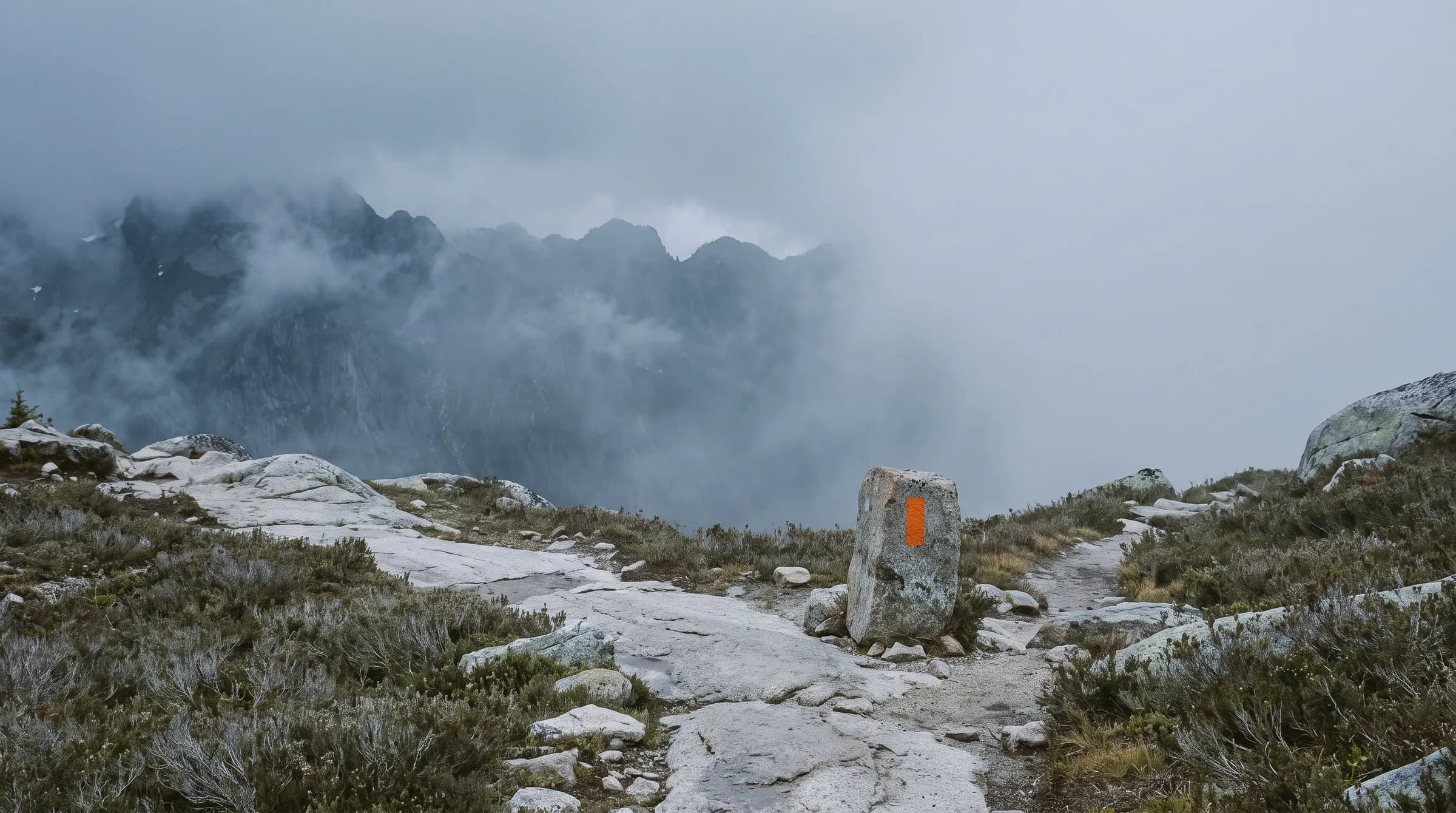 A misty granite mountain trail at a fork, with a single orange trail-marker blaze painted on a standing boulder