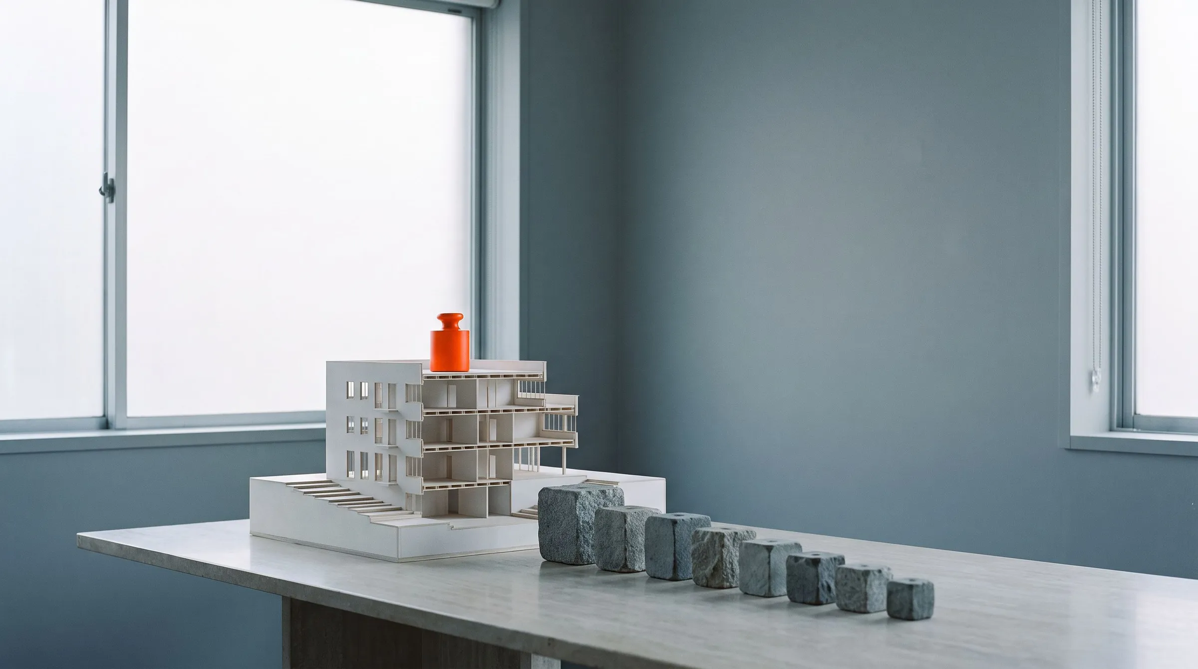 An architect’s cross-section model of a building on a stone table, with a row of grey stone weights beside it and one orange weight on its roof