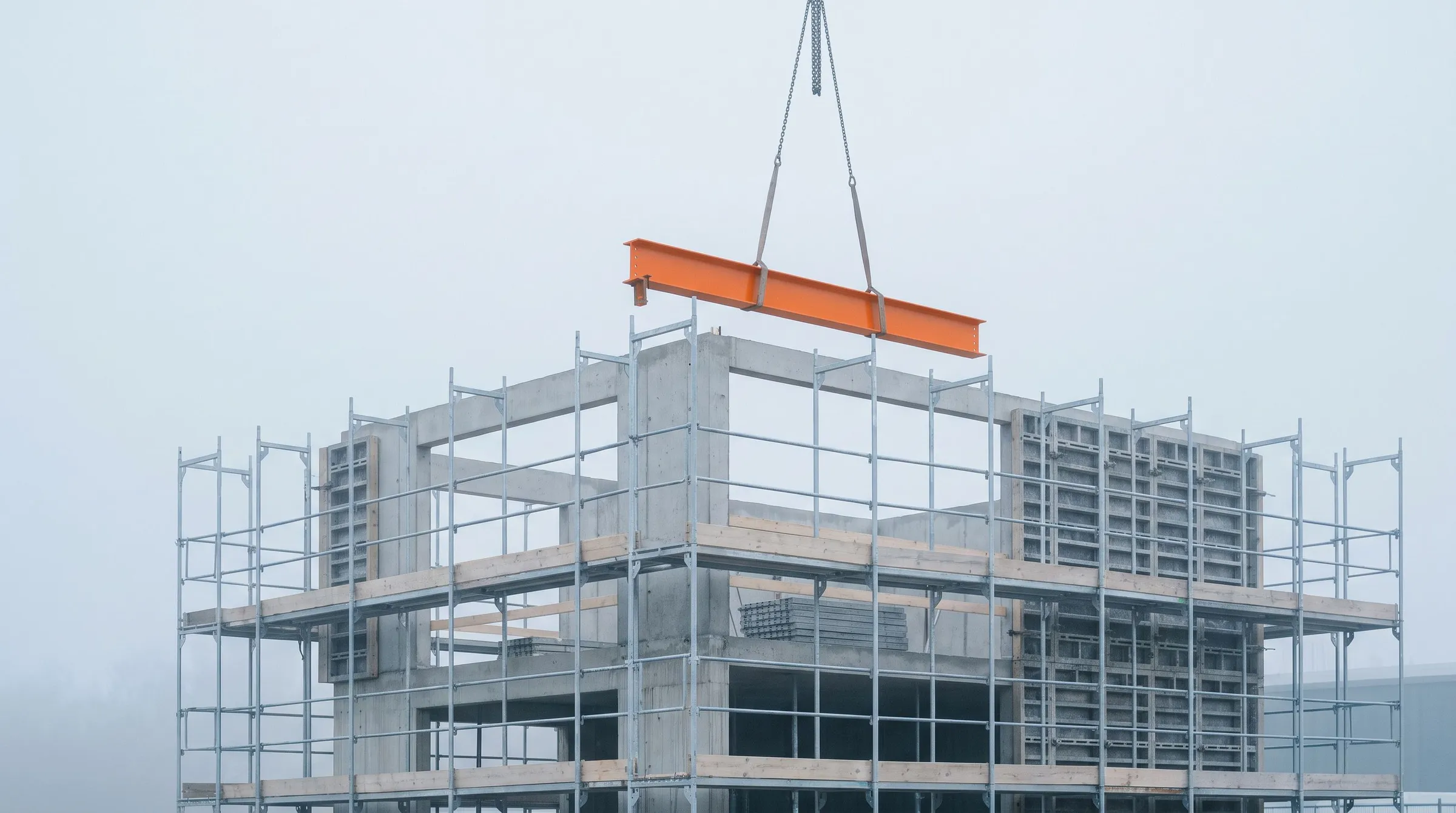 Galvanized scaffolding around a raw concrete frame in the fog, with a single orange steel beam being lowered into place