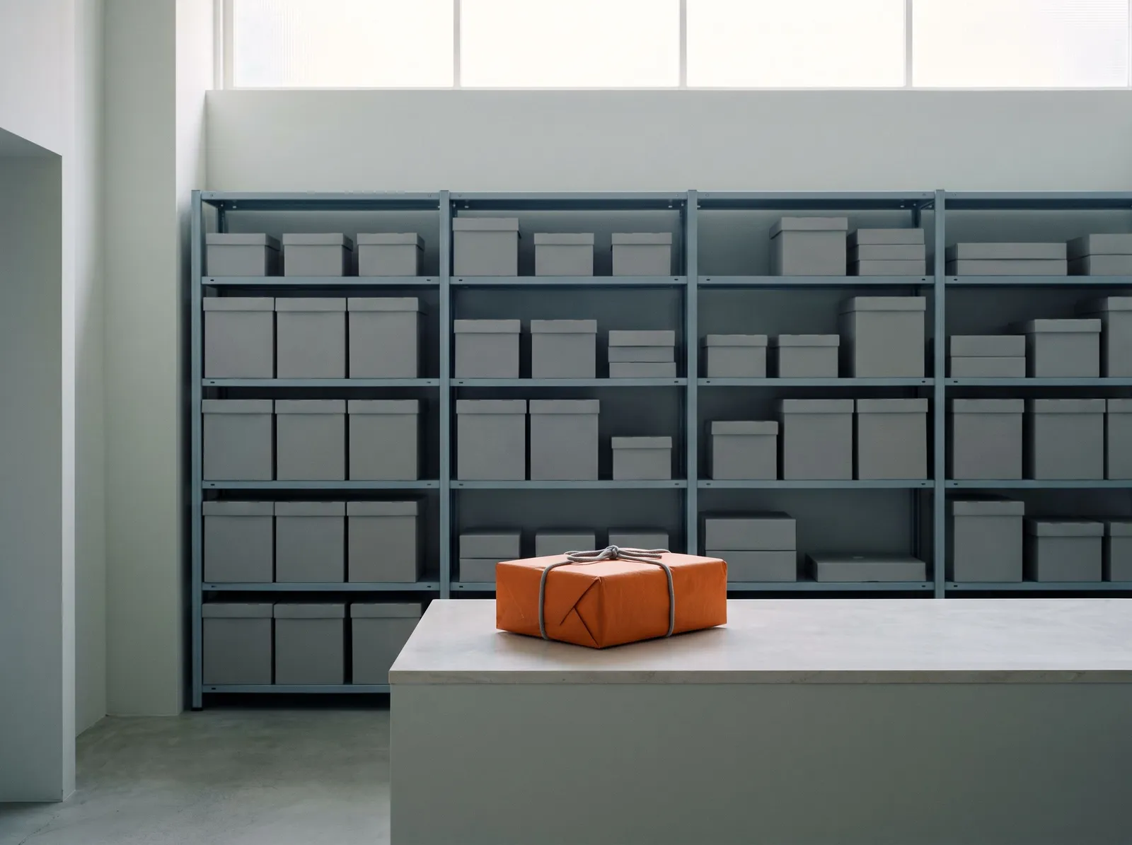 A tidy stockroom with grey unmarked boxes on steel shelving and a single parcel wrapped in orange paper on the dispatch counter