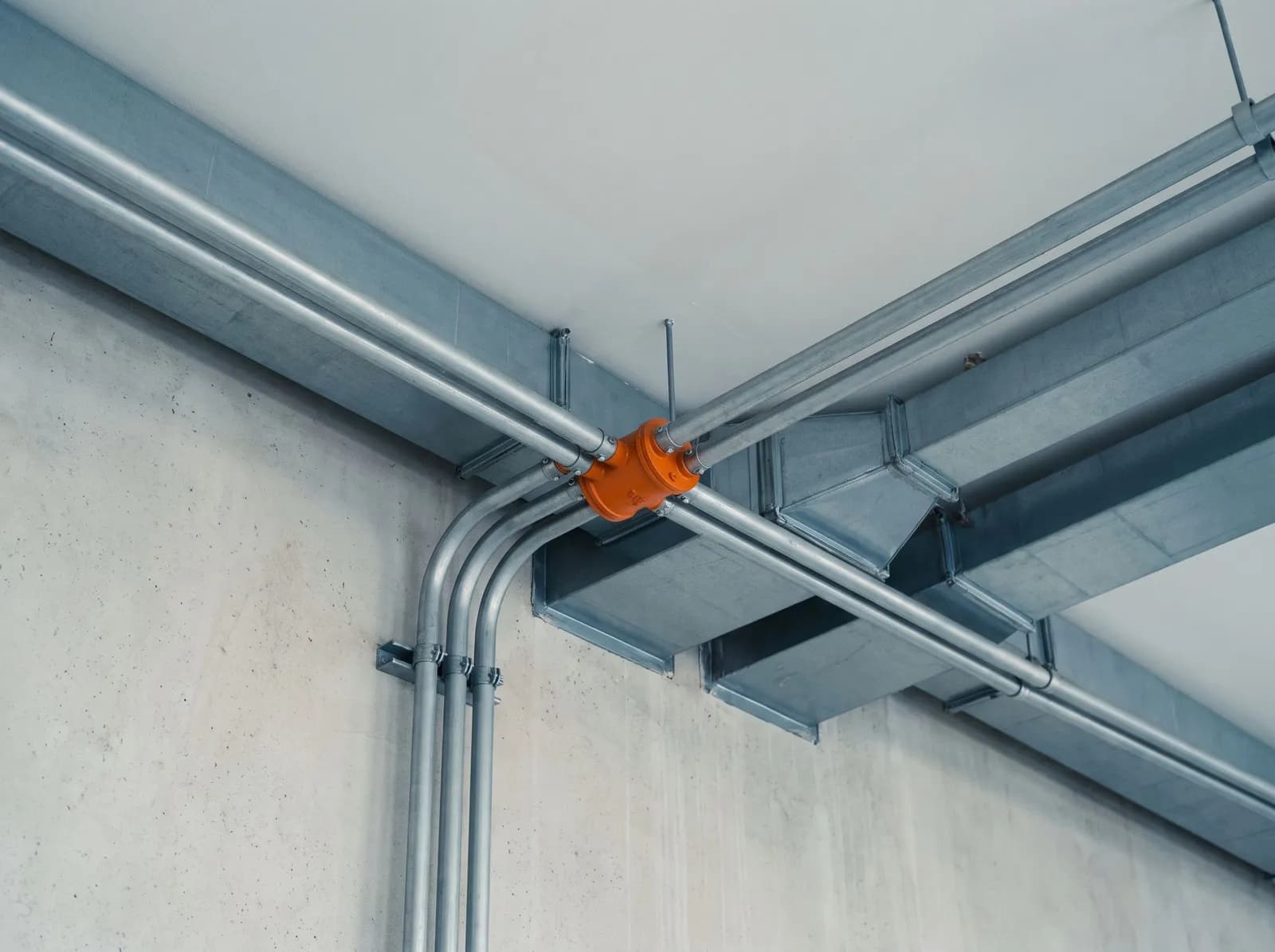 Grey pipes and ducts running along a concrete wall and ceiling, meeting at a single orange coupling