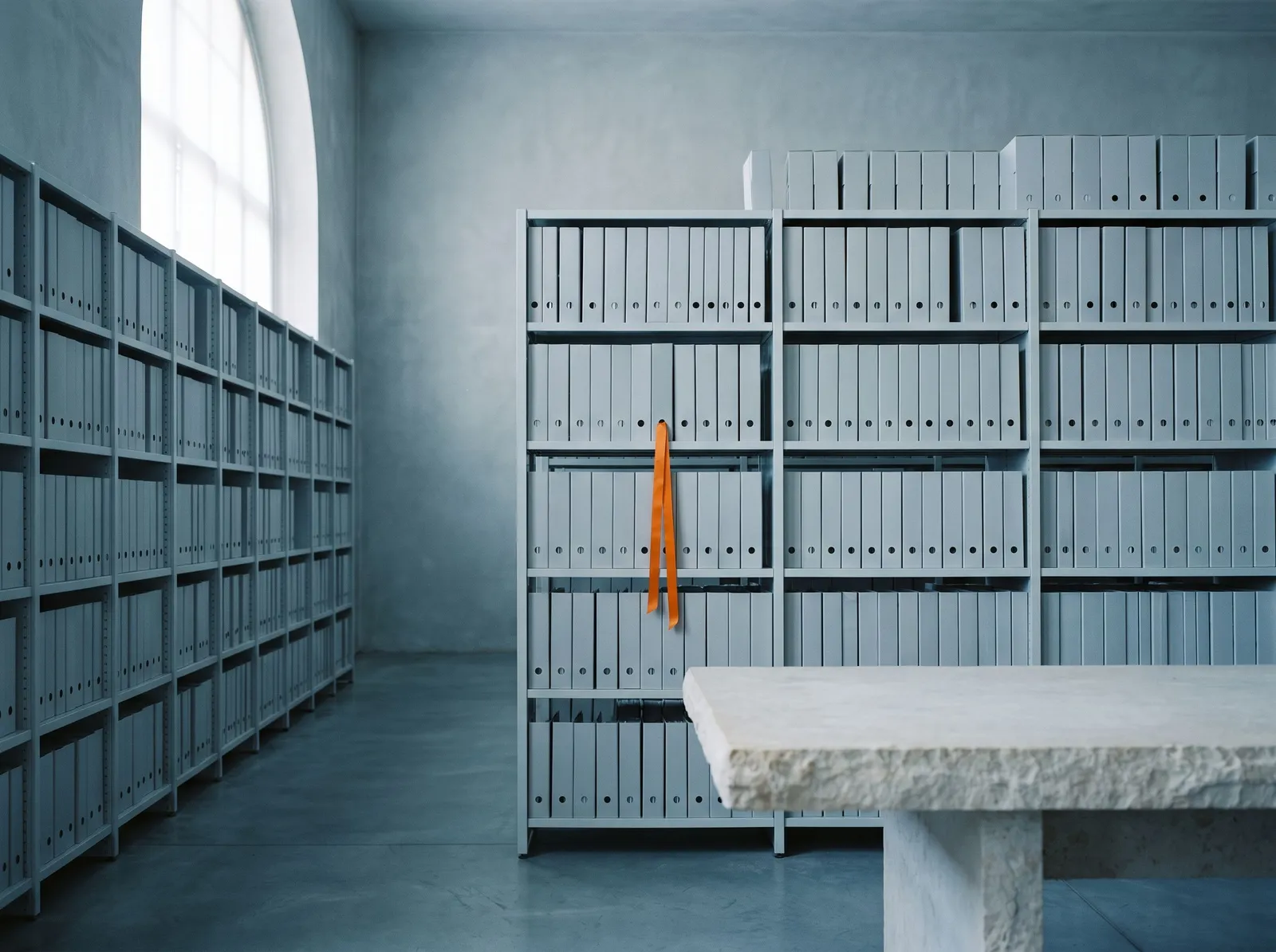 A quiet archive of grey binders on long shelves, with one orange ribbon marking the page that holds the answer