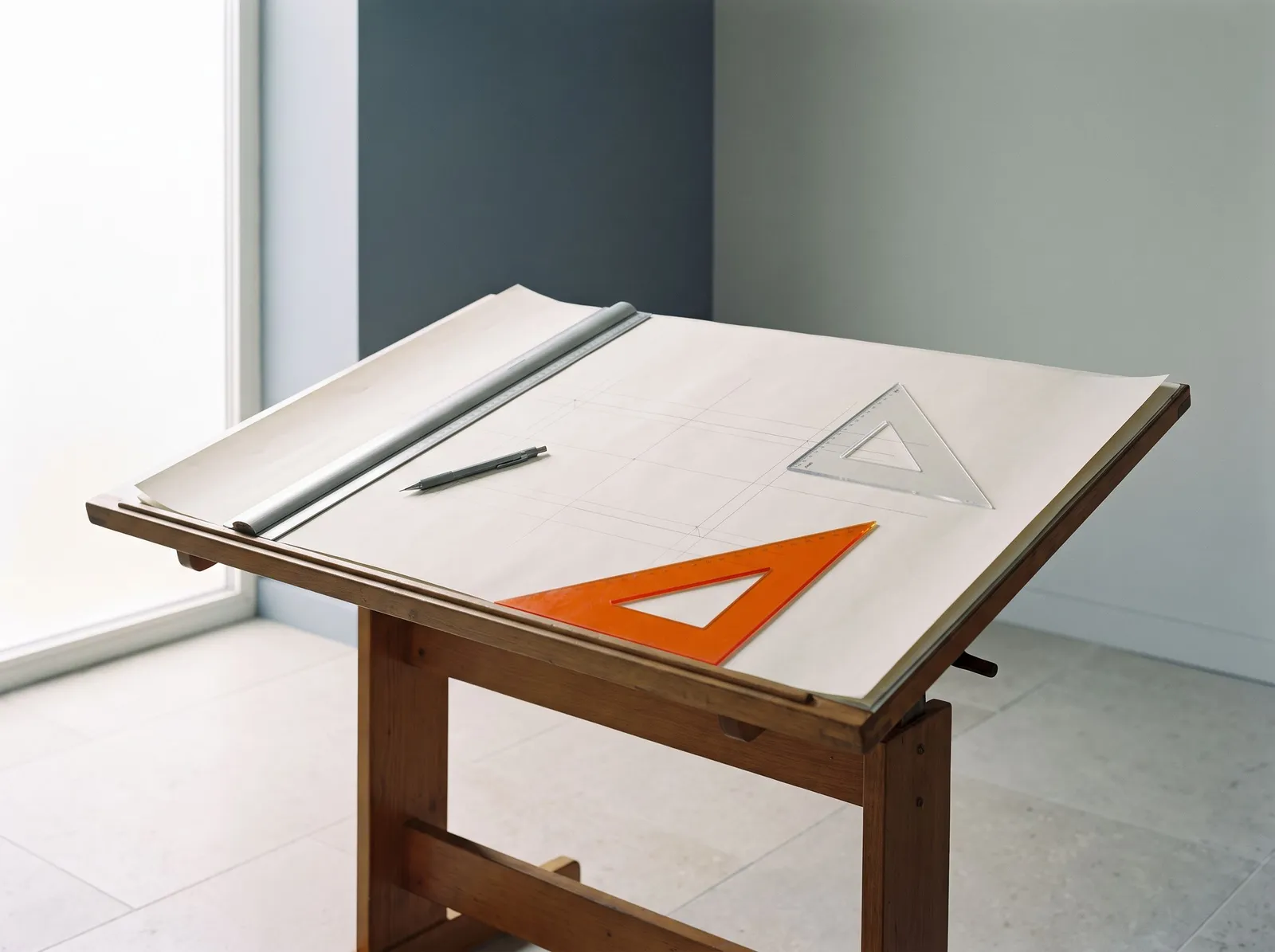 A wooden drafting table with blank paper, a steel parallel ruler, a clear set square and one orange set square, in soft window light